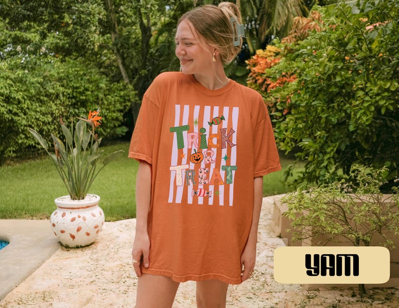 May include: An orange short-sleeved t-shirt featuring a graphic print with white vertical stripes and the text "TRICK TREAT" in colorful, patterned letters. The design includes small illustrations of a pumpkin, candy, and stars.
