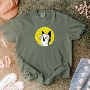 May include: An olive green short-sleeve t-shirt featuring a circular graphic on the chest. The graphic depicts a black and white illustration of a surprised cat with wide eyes and an open mouth, set against a solid, bright yellow circular background.
