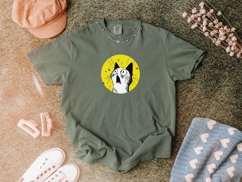 May include: An olive green short-sleeve t-shirt featuring a circular graphic on the chest. The graphic depicts a black and white illustration of a surprised cat with wide eyes and an open mouth, set against a solid, bright yellow circular background.