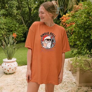 May include: An orange short-sleeve t-shirt featuring a graphic of Santa Claus wearing sunglasses and making a rock-and-roll hand gesture. The design includes a black and white checkered circle, red accents, and the text JINGLE BELL ROCK.
