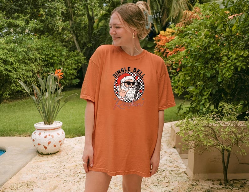 May include: An orange short-sleeve t-shirt featuring a graphic of Santa Claus wearing sunglasses and making a rock-and-roll hand gesture. The design includes a black and white checkered circle, red accents, and the text JINGLE BELL ROCK.