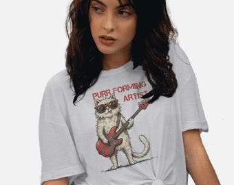 May include: A white short-sleeved t-shirt featuring a graphic illustration of a white cat wearing dark sunglasses and playing a red bass guitar. The text above the cat reads "PURR FORMING ARTIST" in red capital letters. The shirt is tied at the waist.