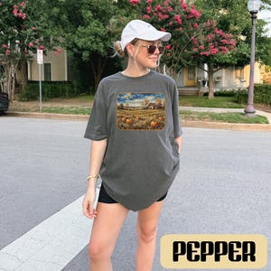 May include: A charcoal gray short-sleeve t-shirt featuring a rectangular graphic print of a rural landscape with a barn and pumpkins in a field. The shirt has a crew neckline and a relaxed fit, paired with a white baseball cap and white sneakers.