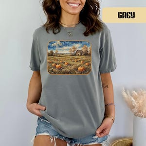 May include: A grey short-sleeve t-shirt featuring a rectangular graphic print of a rural landscape with a barn, a field of orange pumpkins, and a swirling blue sky. The text GREY is printed in a black rectangular box in the upper right corner.