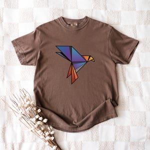 Puede incluir: Una camiseta marr&oacute;n de manga corta con un gr&aacute;fico de p&aacute;jaro de origami geom&eacute;trico en el pecho. El dise&ntilde;o del p&aacute;jaro est&aacute; compuesto por formas angulares en tonos azul, morado y naranja con contornos negros marcados, centrado en la prenda.