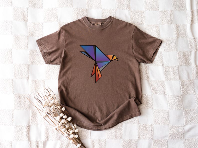 Puede incluir: Una camiseta marr&oacute;n de manga corta con un gr&aacute;fico de p&aacute;jaro de origami geom&eacute;trico en el pecho. El dise&ntilde;o del p&aacute;jaro est&aacute; compuesto por formas angulares en tonos azul, morado y naranja con contornos negros marcados, centrado en la prenda.