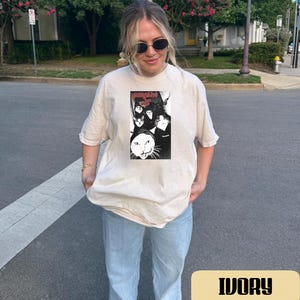 May include: An ivory short-sleeve t-shirt featuring a rectangular black and white graphic print on the chest. The graphic displays multiple cat faces and the text "gangster cat" in a red stylized font. The shirt has a crew neckline and a loose fit.