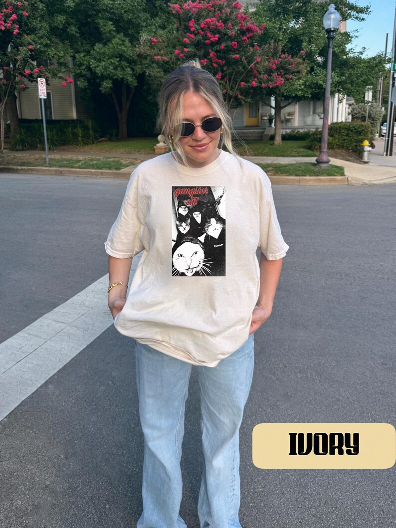 May include: An ivory short-sleeve t-shirt featuring a rectangular black and white graphic print on the chest. The graphic displays multiple cat faces and the text "gangster cat" in a red stylized font. The shirt has a crew neckline and a loose fit.