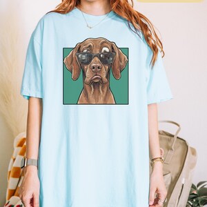 May include: A light blue t-shirt featuring a graphic print of a brown dog wearing black sunglasses. The dog has one eye looking in a different direction and a sad expression. The graphic is set against a solid green square background.