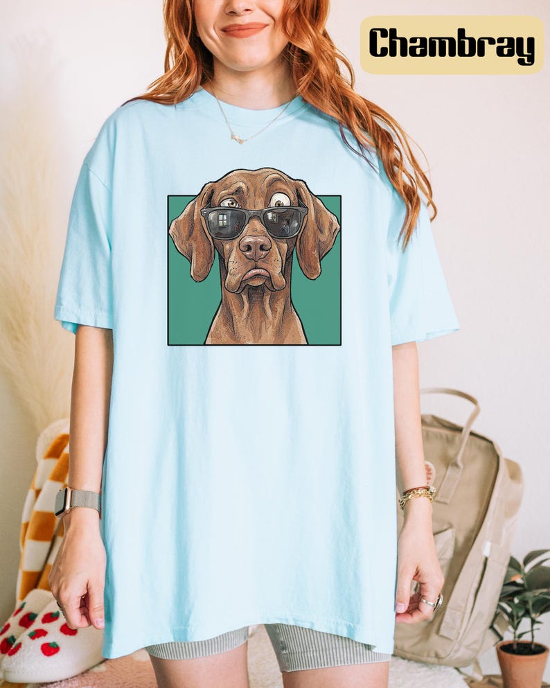 May include: A light blue t-shirt featuring a graphic print of a brown dog wearing black sunglasses. The dog has one eye looking in a different direction and a sad expression. The graphic is set against a solid green square background.