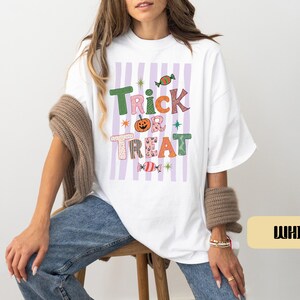 May include: A white t-shirt featuring a graphic print with the text Trick or Treat in colorful, patterned letters. The design includes a central orange pumpkin, two wrapped candies, and small star accents over a background of vertical light purple stripes.