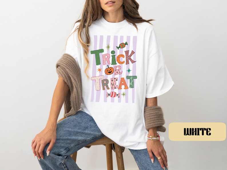 May include: A white t-shirt featuring a graphic print with the text Trick or Treat in colorful, patterned letters. The design includes a central orange pumpkin, two wrapped candies, and small star accents over a background of vertical light purple stripes.