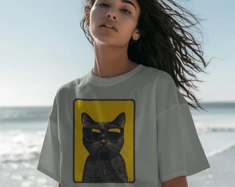 May include: A light gray t-shirt featuring a rectangular graphic print of a black cat wearing dark sunglasses against a solid yellow background. The cat is centered within the frame, displaying detailed fur texture and a stoic expression.