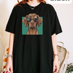 May include: A black short-sleeved t-shirt featuring a graphic print of a brown dog wearing black sunglasses. The dog has a surprised expression with wide eyes and is set against a solid teal square background. The word Black is printed in white.