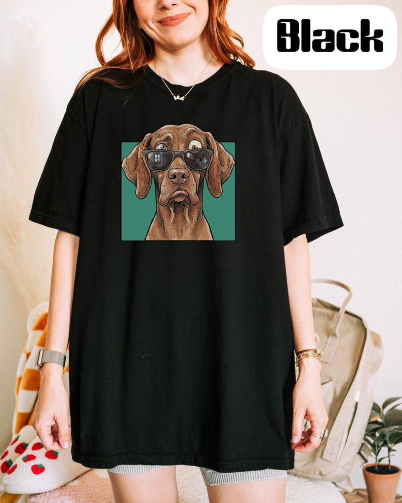 May include: A black short-sleeved t-shirt featuring a graphic print of a brown dog wearing black sunglasses. The dog has a surprised expression with wide eyes and is set against a solid teal square background. The word Black is printed in white.