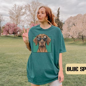 May include: A dark teal short-sleeved t-shirt featuring a graphic print of a brown dog wearing black sunglasses inside a square frame. The shirt has a crew neckline and a relaxed fit, with the text BLUE SPRUCE displayed in a rectangular box.