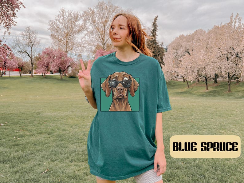 May include: A dark teal short-sleeved t-shirt featuring a graphic print of a brown dog wearing black sunglasses inside a square frame. The shirt has a crew neckline and a relaxed fit, with the text BLUE SPRUCE displayed in a rectangular box.