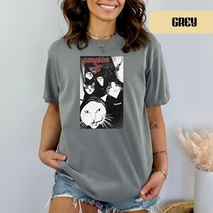 May include: A grey short-sleeve t-shirt featuring a black and white graphic print of multiple cats with the text "gangster cat" in red lettering above the image. The shirt has a crew neckline and a relaxed fit, showcasing a vintage-style design.