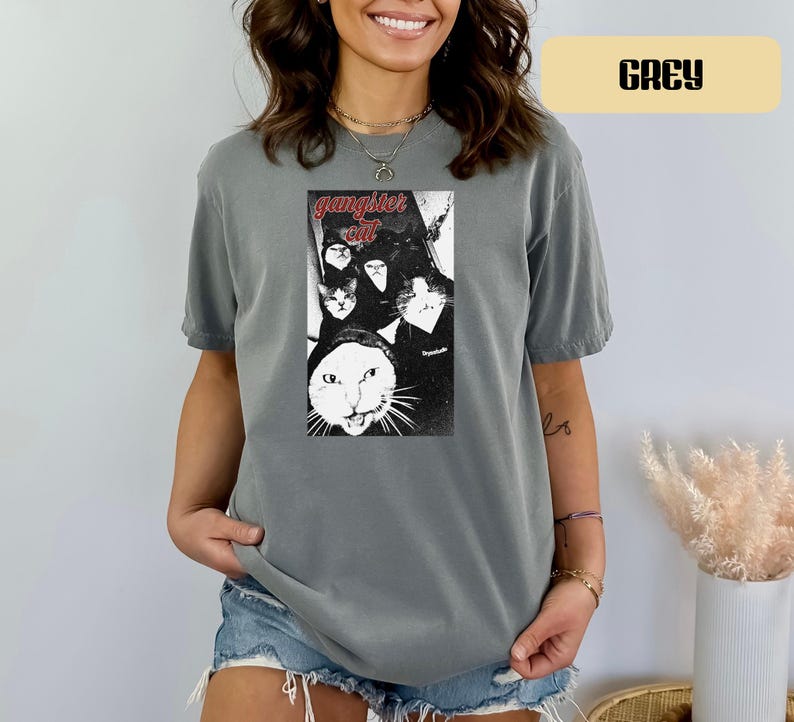 May include: A grey short-sleeve t-shirt featuring a black and white graphic print of multiple cats with the text "gangster cat" in red lettering above the image. The shirt has a crew neckline and a relaxed fit, showcasing a vintage-style design.