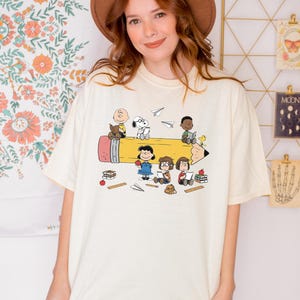 May include: An ivory short-sleeve t-shirt featuring a graphic print of Peanuts characters including Charlie Brown, Snoopy, and others interacting with a large yellow pencil, books, and apples. The word Ivory is printed in black at the top right corner.