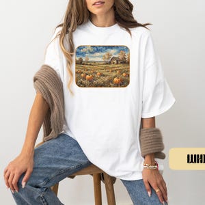 May include: A white short-sleeved t-shirt featuring a rectangular graphic print of a rural landscape with a pumpkin patch, a barn, and trees under a cloudy sky. The shirt has a loose fit and is paired with blue denim jeans and a brown knit sweater.