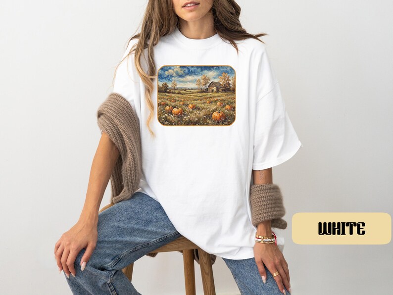 May include: A white short-sleeved t-shirt featuring a rectangular graphic print of a rural landscape with a pumpkin patch, a barn, and trees under a cloudy sky. The shirt has a loose fit and is paired with blue denim jeans and a brown knit sweater.