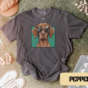 May include: A charcoal grey short-sleeve t-shirt featuring a graphic print of a brown dog wearing black sunglasses. The dog is centred within a teal square frame. The shirt has a crew neckline and a relaxed fit, with the word PEPPER printed below.