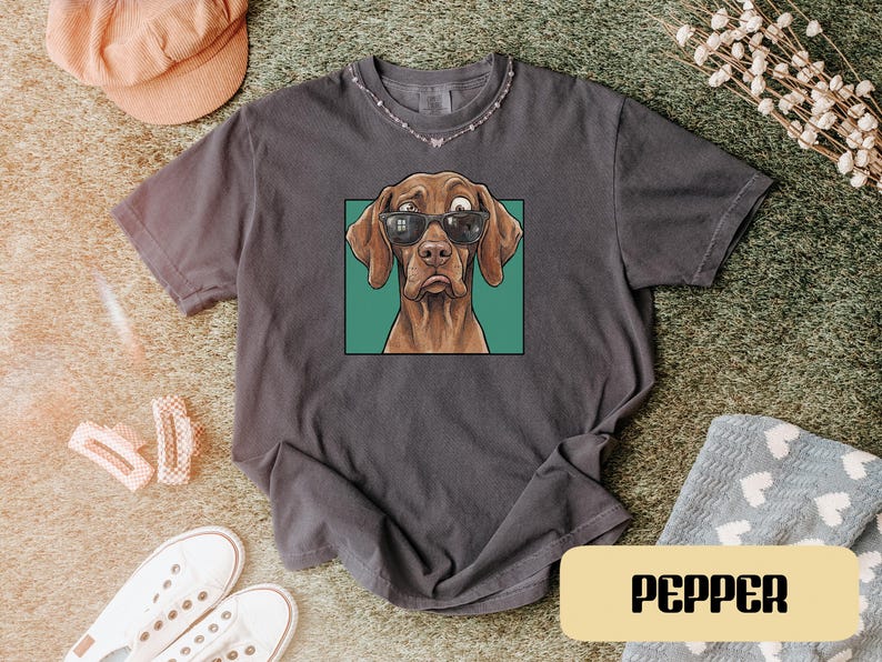 May include: A charcoal grey short-sleeve t-shirt featuring a graphic print of a brown dog wearing black sunglasses. The dog is centred within a teal square frame. The shirt has a crew neckline and a relaxed fit, with the word PEPPER printed below.