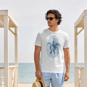 May include: A white short-sleeved t-shirt featuring a blue graphic print of a sea turtle centered within a fingerprint pattern. The shirt is paired with light blue shorts and a straw hat with a black band, worn with dark sunglasses.
