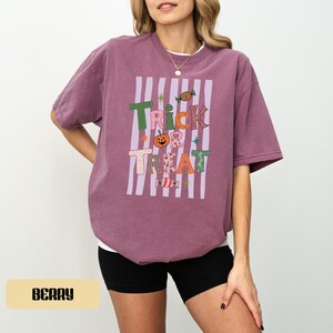 May include: A berry-colored short-sleeve t-shirt featuring a graphic print with the text Trick or Treat. The design includes a pumpkin, candy, and star icons over vertical light purple stripes. The shirt is paired with black athletic shorts.