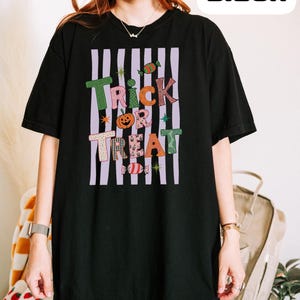 May include: A black short-sleeve t-shirt featuring a graphic design with vertical light purple stripes and the text Trick or Treat in colorful, patterned letters. The design includes small illustrations of a pumpkin and candies.