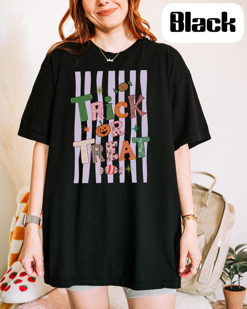 May include: A black short-sleeve t-shirt featuring a graphic design with vertical light purple stripes and the text Trick or Treat in colorful, patterned letters. The design includes small illustrations of a pumpkin and candies.