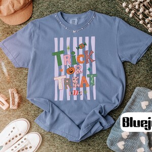 May include: A blue short-sleeve t-shirt featuring a graphic print with the text "TRICK TREAT" in colorful, patterned letters. The design includes a pumpkin, candy illustrations, and vertical light purple stripes on a blue background.
