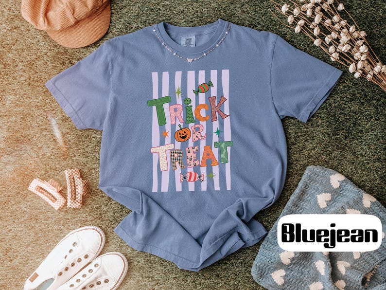 May include: A blue short-sleeve t-shirt featuring a graphic print with the text "TRICK TREAT" in colorful, patterned letters. The design includes a pumpkin, candy illustrations, and vertical light purple stripes on a blue background.