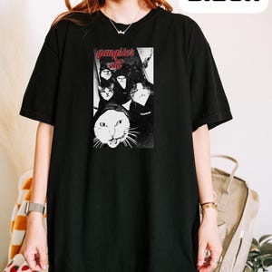 May include: A black short-sleeved t-shirt featuring a graphic print of multiple cats with the red text "gangster cat" above them. The design is centered on the chest area of the shirt, which also includes the word "Black" in a white box.