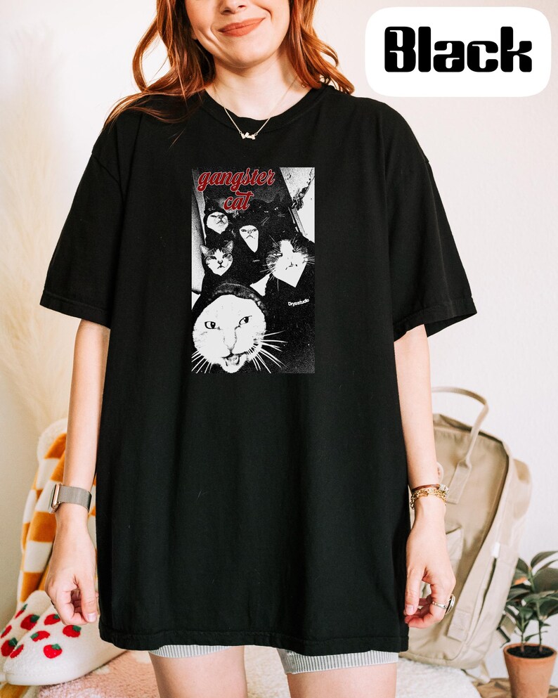 May include: A black short-sleeved t-shirt featuring a graphic print of multiple cats with the red text "gangster cat" above them. The design is centered on the chest area of the shirt, which also includes the word "Black" in a white box.