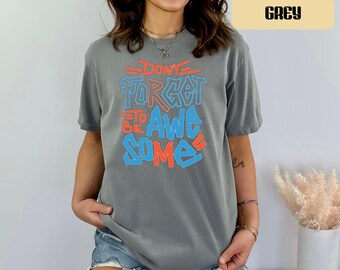 May include: A grey short-sleeve t-shirt featuring the text "DON'T FORGET TO BE AWESOME" printed in a layered style with blue and orange lettering. The shirt has a crew neckline and a relaxed fit, paired with blue denim shorts with frayed hems.