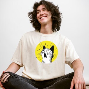 May include: A cream-colored short-sleeve t-shirt featuring a circular graphic on the chest. The graphic depicts a black and white line-art illustration of a cat with wide, surprised eyes and an open mouth, set against a solid bright yellow background.