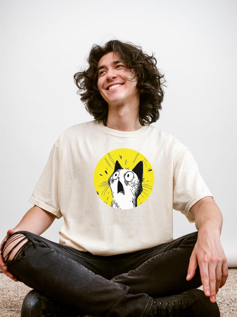 May include: A cream-colored short-sleeve t-shirt featuring a circular graphic on the chest. The graphic depicts a black and white line-art illustration of a cat with wide, surprised eyes and an open mouth, set against a solid bright yellow background.