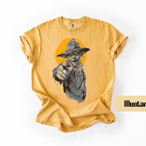 May include: A mustard yellow short-sleeve t-shirt featuring a graphic illustration of a scarecrow wearing a hat and pointing forward. The scarecrow is set against a solid orange circle. The word "Mustard" is printed in a small box on the side.
