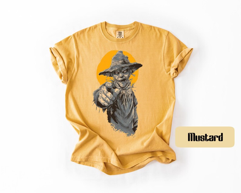 May include: A mustard yellow short-sleeve t-shirt featuring a graphic illustration of a scarecrow wearing a hat and pointing forward. The scarecrow is set against a solid orange circle. The word "Mustard" is printed in a small box on the side.
