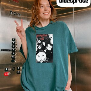 May include: A dark teal short-sleeve t-shirt featuring a rectangular graphic print of multiple cats with the text "gangster cat" in red lettering at the top. The shirt has a crew neckline and a loose fit, showcasing a black and white cat illustration.