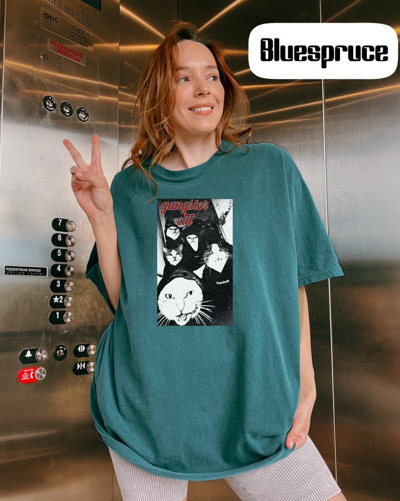 May include: A dark teal short-sleeve t-shirt featuring a rectangular graphic print of multiple cats with the text "gangster cat" in red lettering at the top. The shirt has a crew neckline and a loose fit, showcasing a black and white cat illustration.