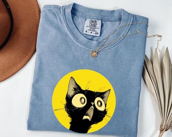 May include: A blue short-sleeved t-shirt featuring a graphic of a black cat with large, wide eyes and an open mouth set against a bright yellow circular background. The shirt has a Comfort Colors brand label at the neck and a crew neckline.