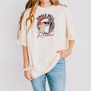 May include: A cream-colored short-sleeve t-shirt featuring a graphic of Santa Claus wearing sunglasses and making a rock-and-roll hand gesture. The design includes a black and white checkered circle, red stars, and the text JINGLE BELL Rockin.