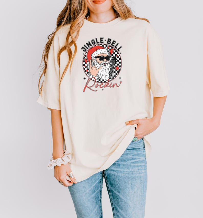 May include: A cream-colored short-sleeve t-shirt featuring a graphic of Santa Claus wearing sunglasses and making a rock-and-roll hand gesture. The design includes a black and white checkered circle, red stars, and the text JINGLE BELL Rockin.