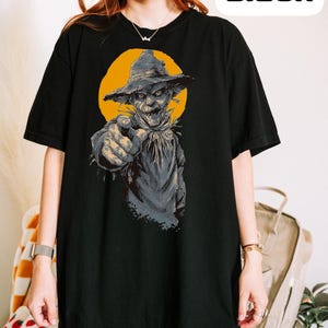 May include: A black short-sleeve t-shirt featuring a graphic print of a scarecrow wearing a hat and pointing forward against a yellow circular background. The word Black is printed in a bold, stylized font in the upper right corner of the image.
