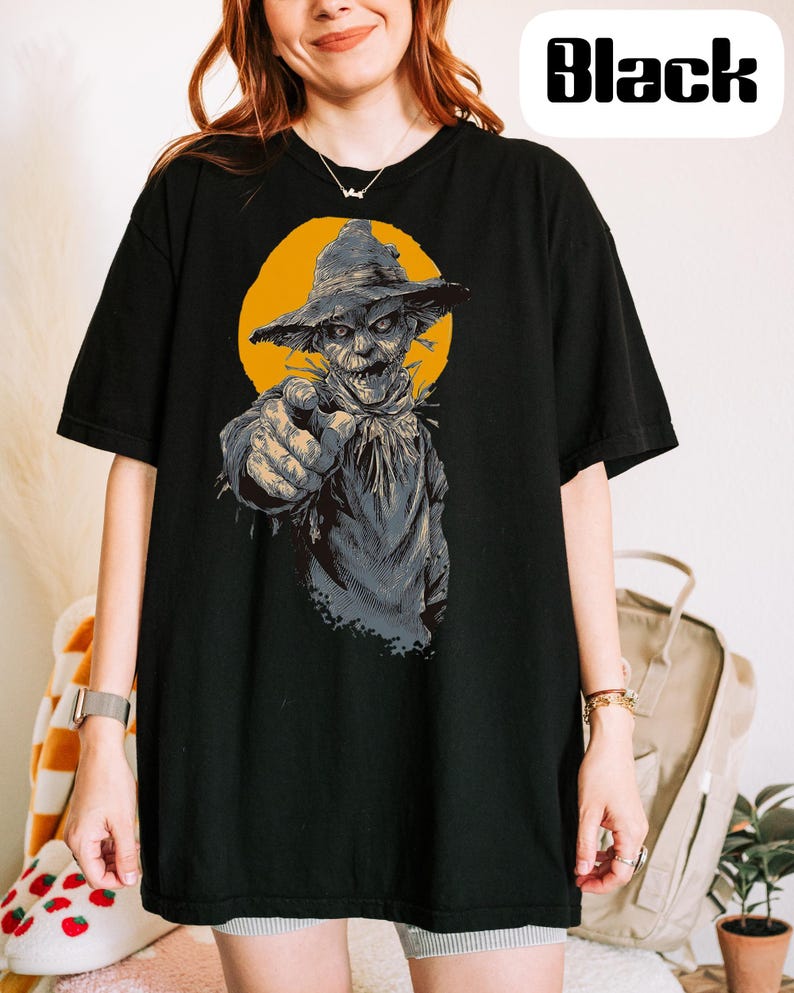 May include: A black short-sleeve t-shirt featuring a graphic print of a scarecrow wearing a hat and pointing forward against a yellow circular background. The word Black is printed in a bold, stylized font in the upper right corner of the image.