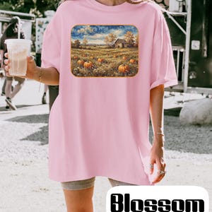May include: A light pink short-sleeved t-shirt featuring a rectangular graphic print of a rural landscape with orange pumpkins in a field, trees, a small barn, and a blue sky with clouds. The shirt has a crew neckline and a loose, relaxed fit.