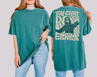 May include: A teal short-sleeve t-shirt featuring a graphic on the back. The design includes a distressed, light-colored illustration of a masked figure with the text "YOU CAN'T PLEASE EVERYONE" in a bold, wavy, retro-style font.